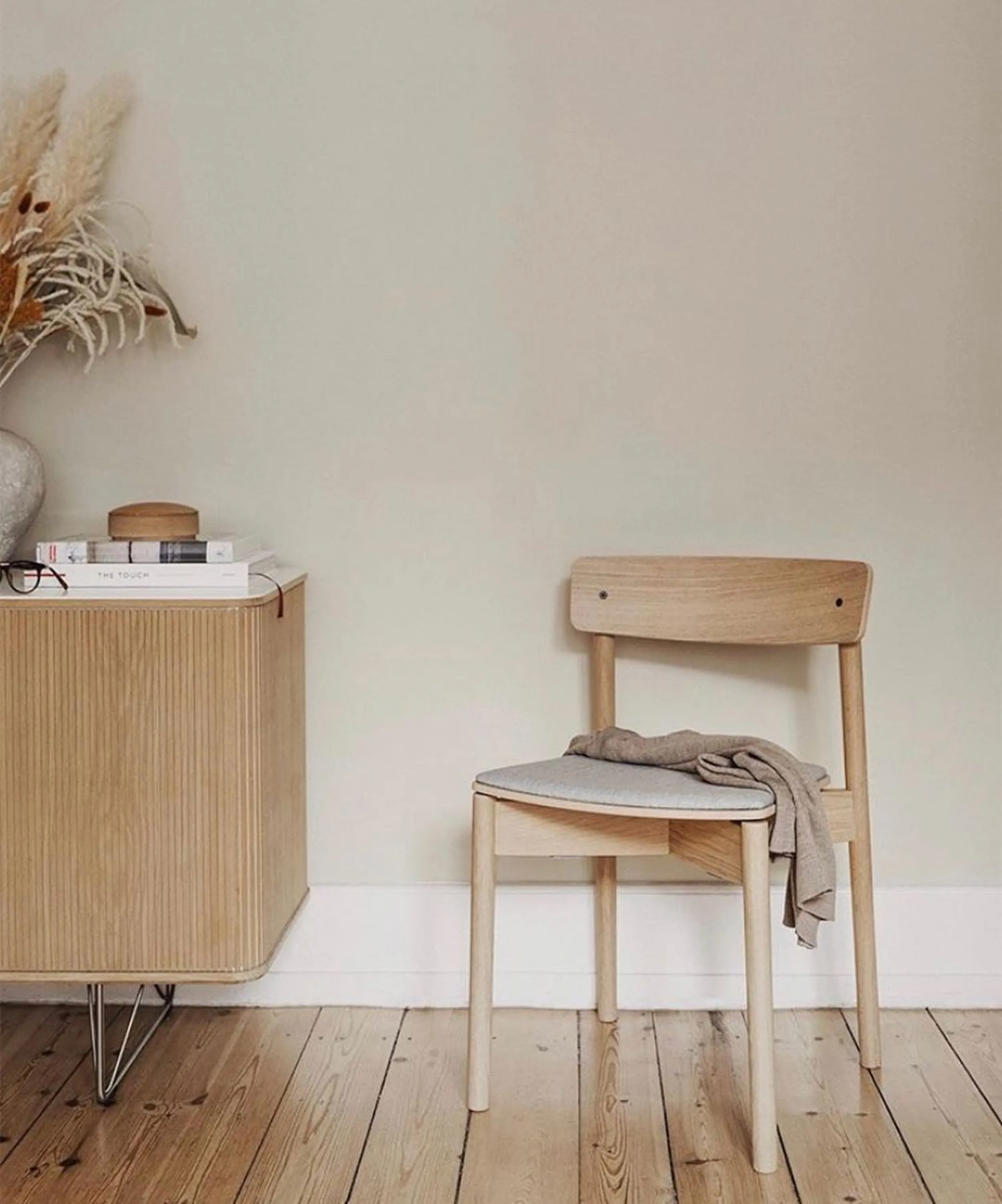 Front view of a sustainable wood chair styled with a blanket on top and a dresser with books and dry flowers on the side.