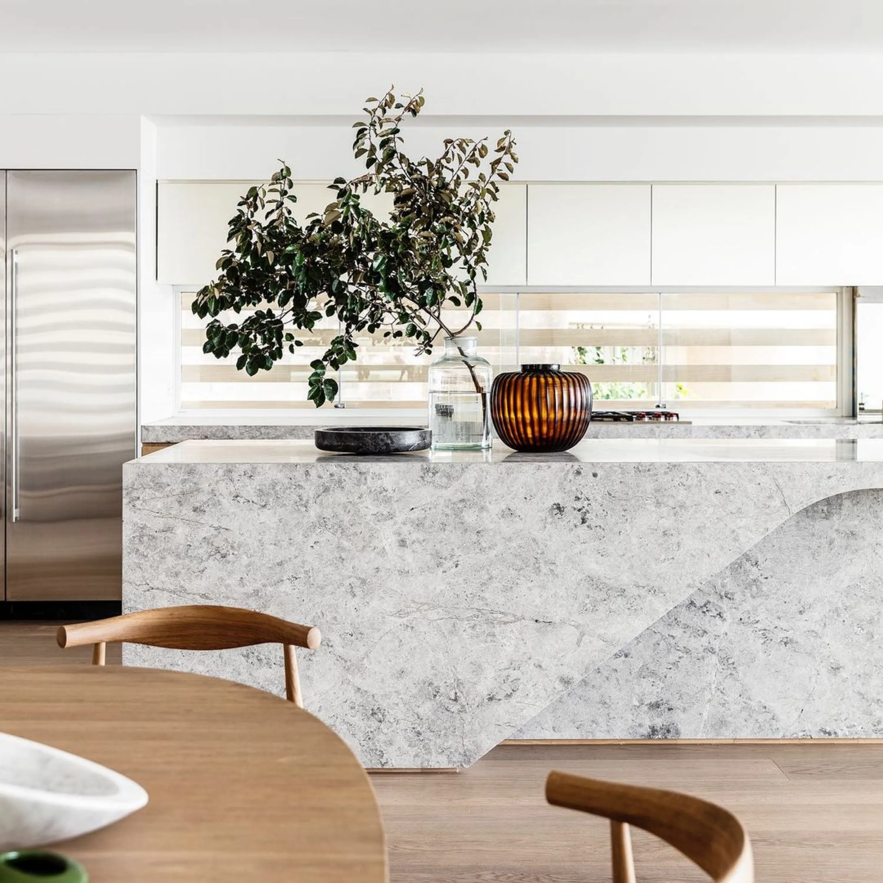 Biophilic dining area looking into an open-plan kitchen, whose stone island and big decorative branches fill the view with natural textures.